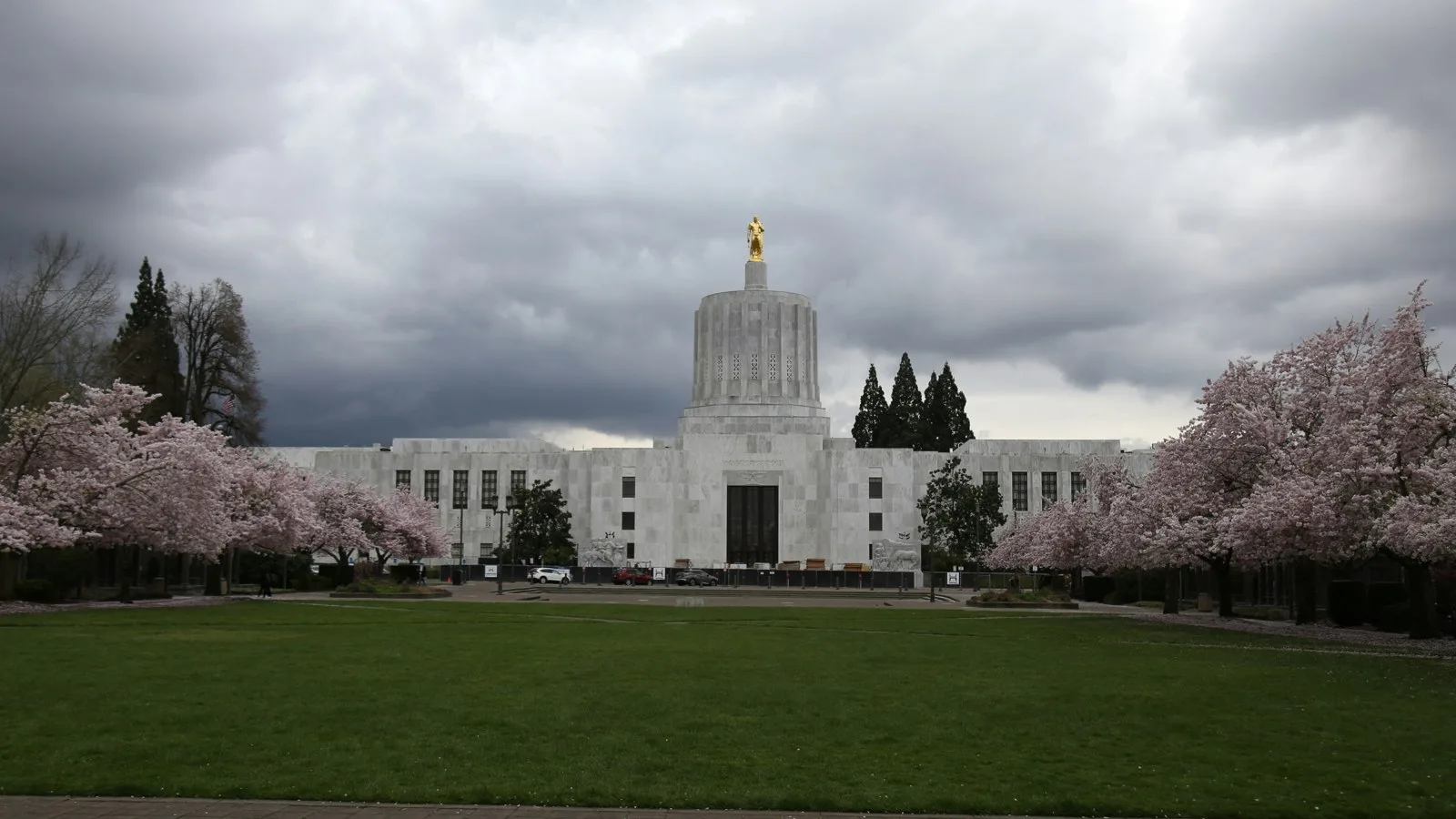 Oregon State Capitol with a gold statue on top, flowering cherry trees on either side and a dark cloudy sky