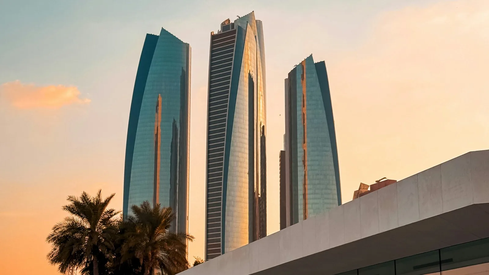 Three curved glass skyscrapers in Abu Dhabi glowing in warm sunset light behind palm trees and the edge of a white building