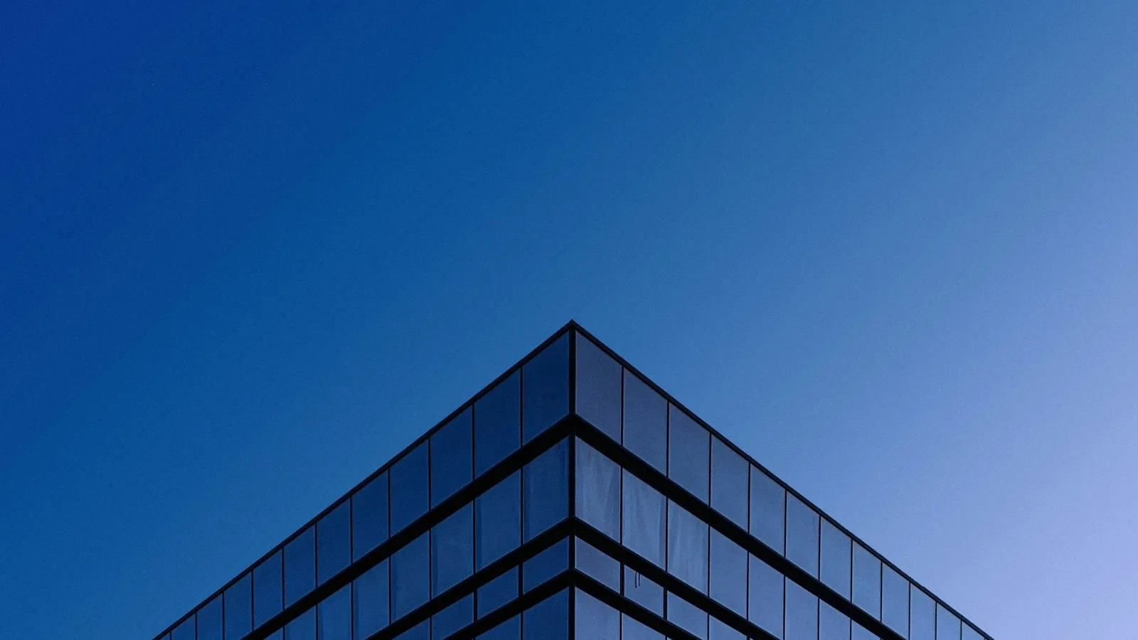 Corner of a glass office building rising against a deep blue sky.