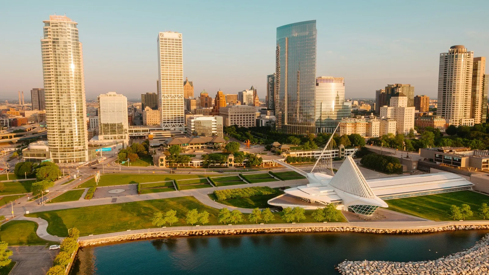 Aerial view of the Milwaukee skyline and lakefront at sunrise, with glass towers, a white lakeside museum building and green parkland