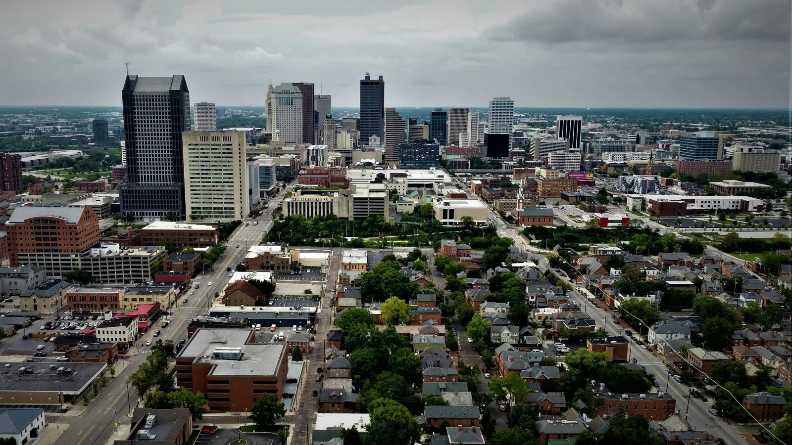 Aerial view of the Columbus skyline with downtown towers rising above brick rowhouses and tree-lined streets under heavy grey clouds