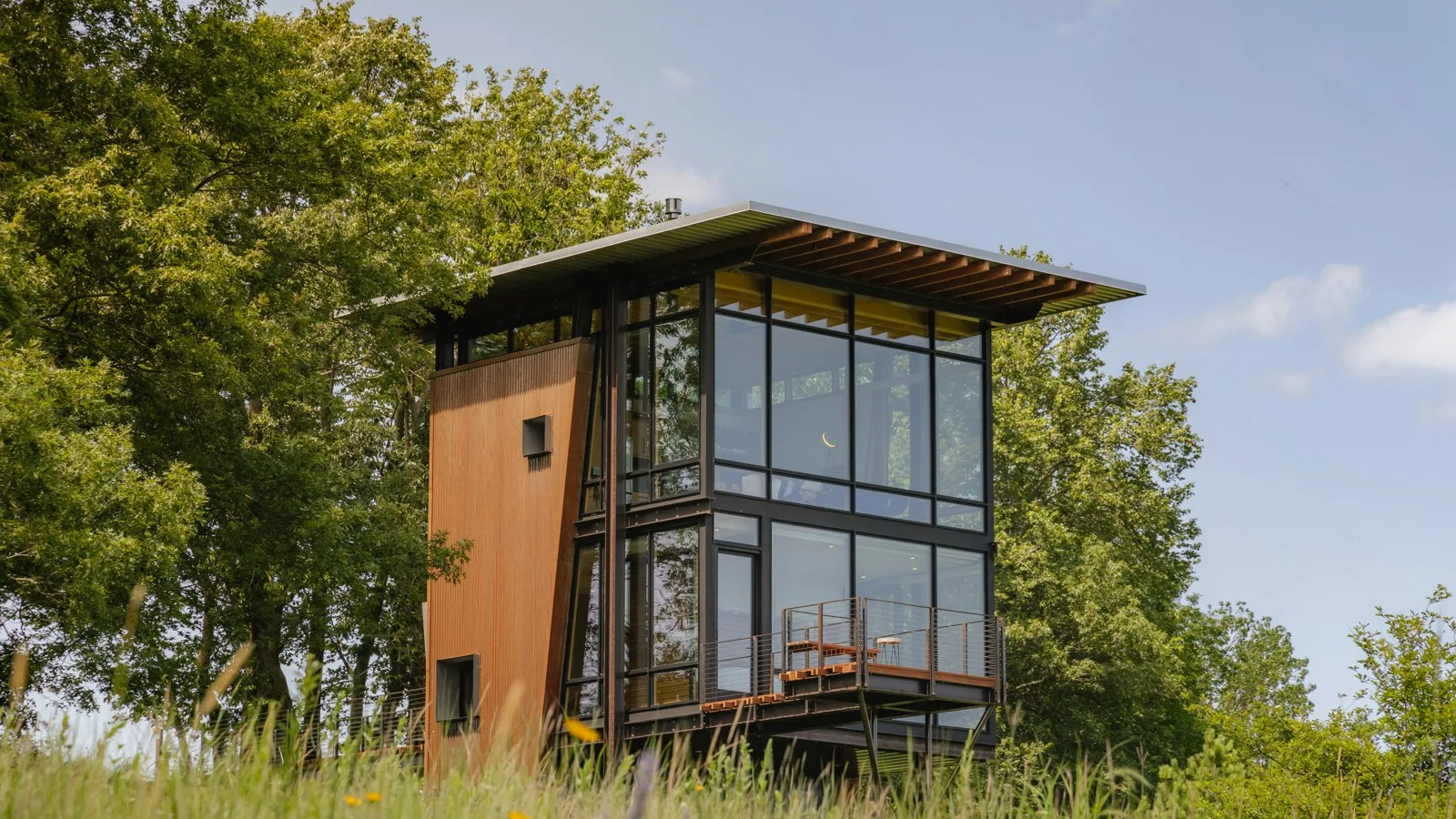 Two-storey glass and timber house with a cantilevered balcony, surrounded by tall trees and long grass under a blue sky