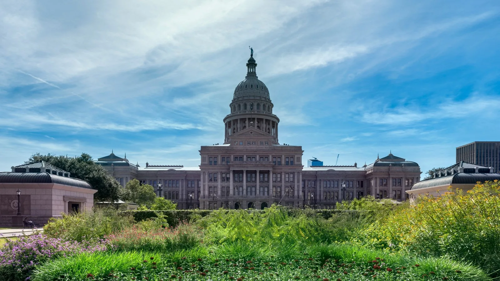 Domed granite state capitol building behind flowerbeds under a blue sky with wispy clouds in Austin, Texas