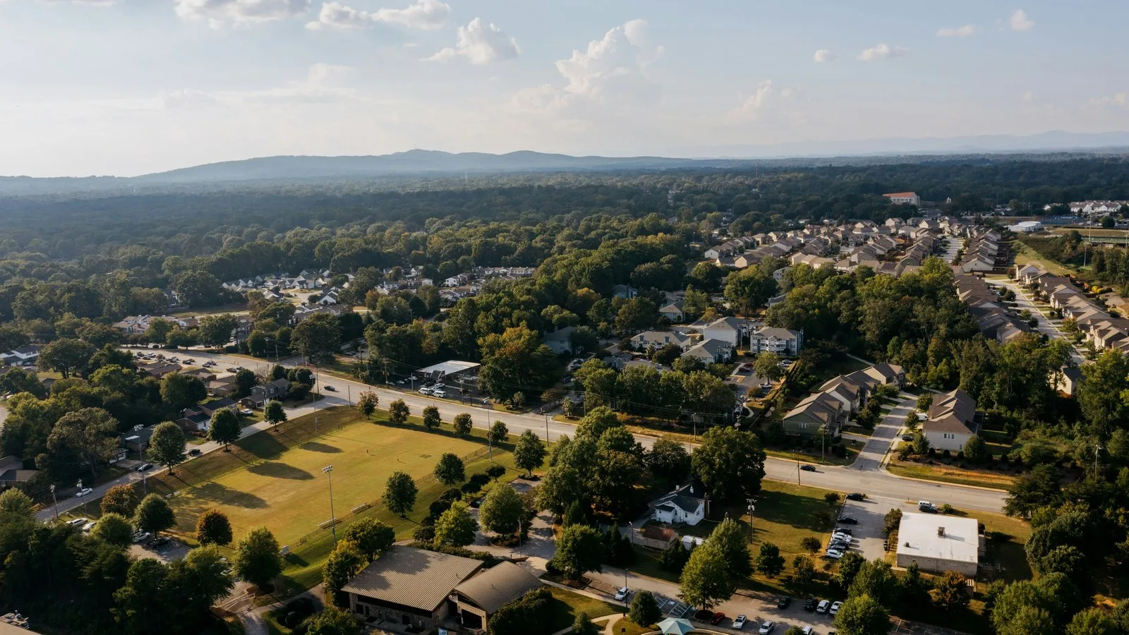 Aerial view of a suburban neighborhood with tree-lined streets, houses, a sports field and a road, with hills in the distance