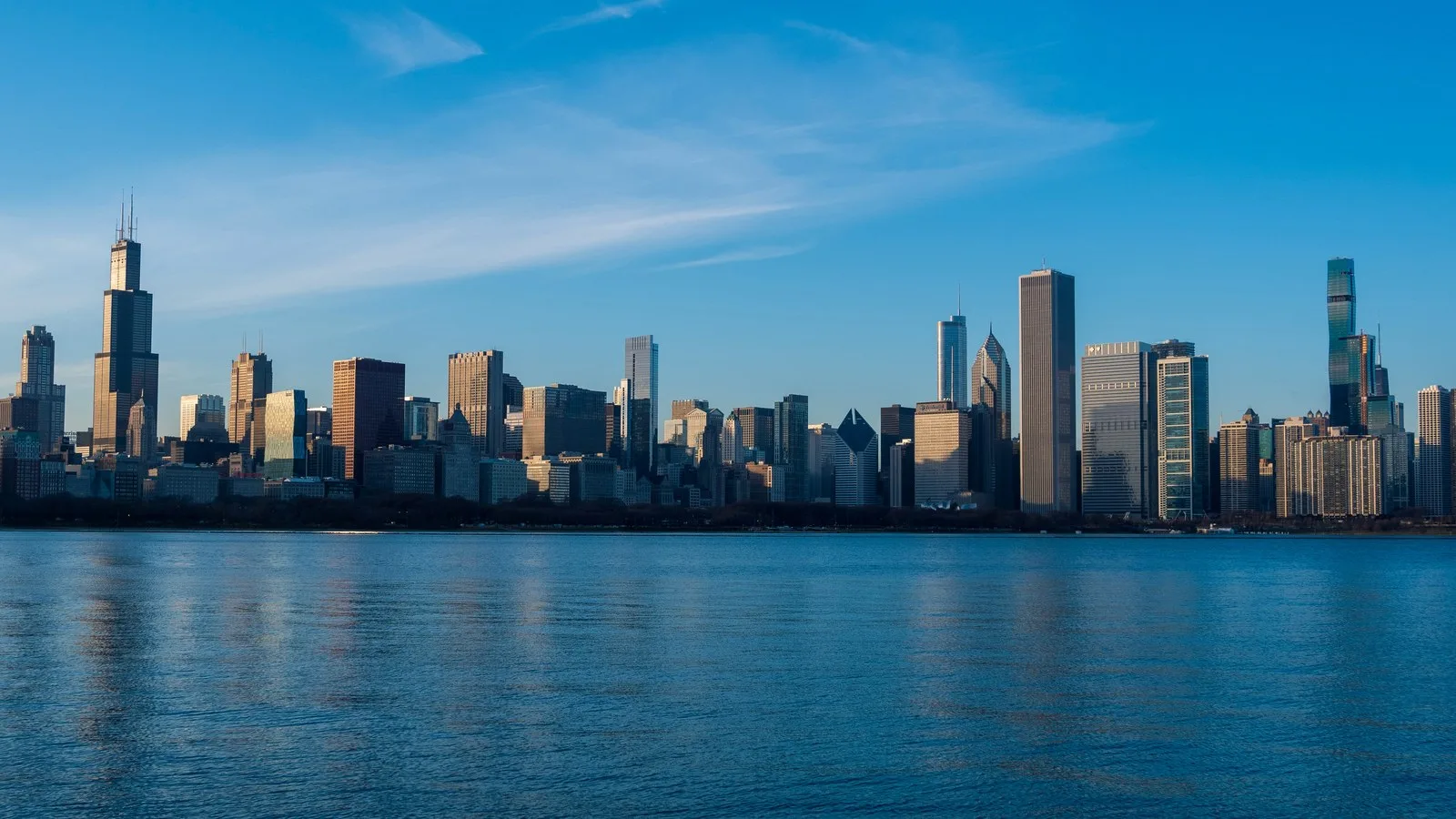 Chicago skyline seen from across the water on a clear afternoon, with tall towers lit by low sun and blue sky above
