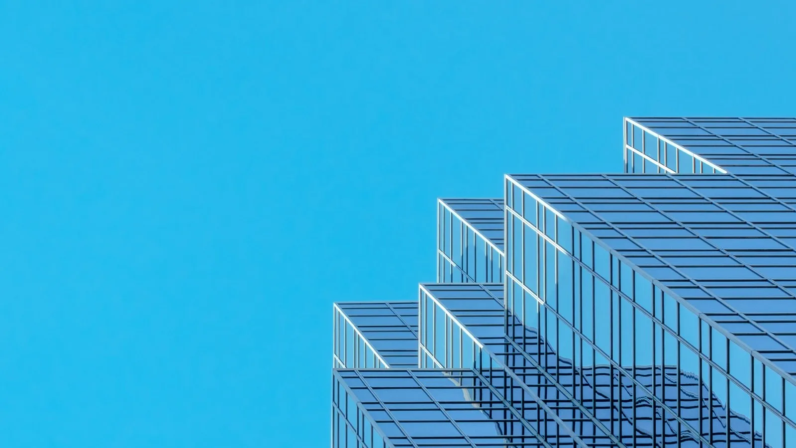 Stepped glass tower facade reflecting blue sky, seen from below against a clear cyan sky