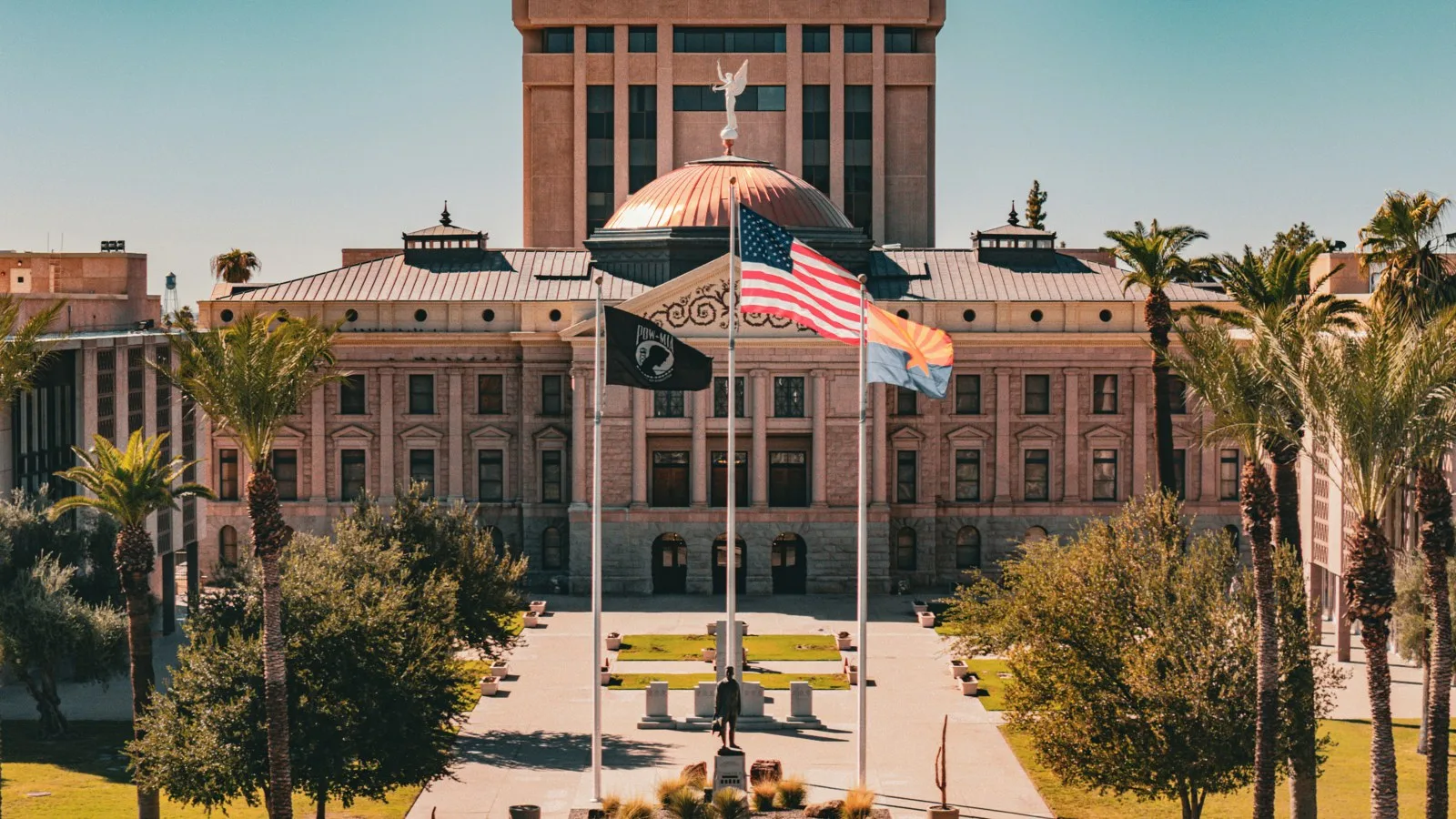 Arizona State Capitol with a copper dome behind palm trees and three flagpoles flying the US and Arizona flags under a clear blue sky