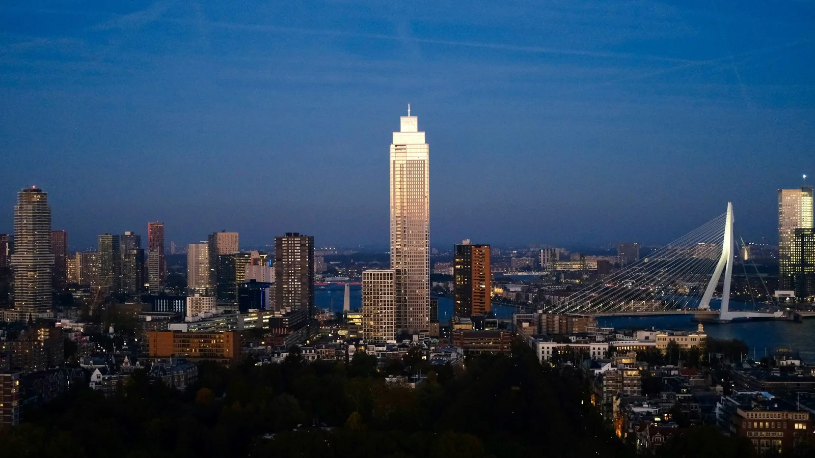 Rotterdam skyline at dusk with a tall tower lit by the last sun beside a white cable-stayed bridge over the river